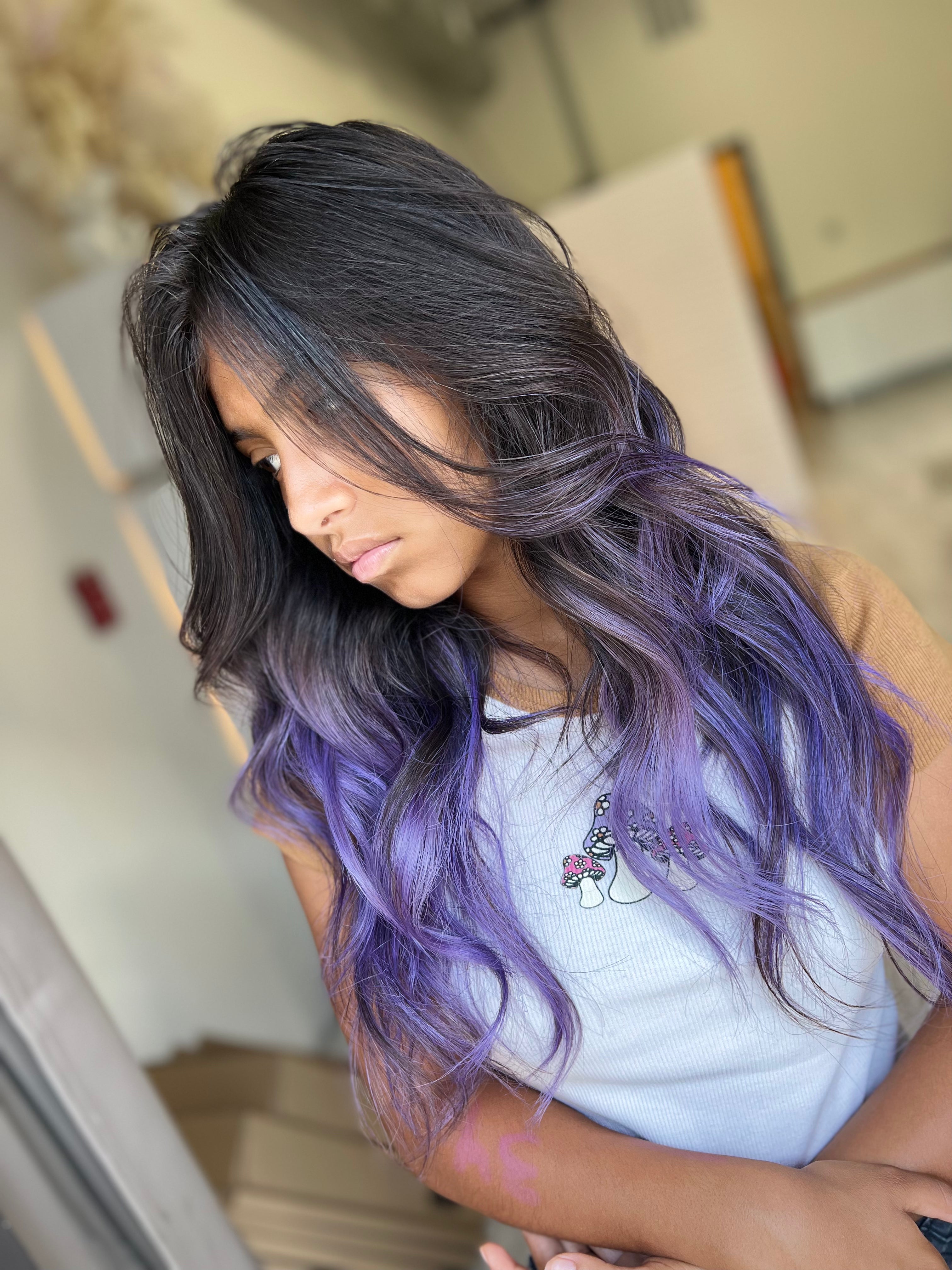 Persona con cabello largo y ondulado que va del negro al morado, vistiendo una camisa azul claro.